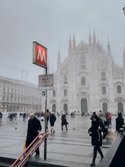 A historical gothic cathedral dominates the background, with mist creating an ethereal atmosphere. People are walking in the large plaza in front of the cathedral, some holding umbrellas. On the left, a red sign with a white 'M' indicates a metro station, and a smaller board below it displays 'Wi-Fi area'. Several pigeons are seen on the ground and a few people are using mobile devices.
