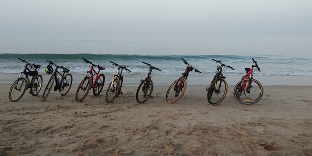 A line of bicycles is parked on sandy beach, facing the ocean. The waves are gently rolling in under a clear sky, creating a serene coastal scene.