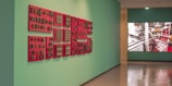 A gallery hallway featuring an art installation on the left with red-framed objects displayed on a teal wall. The installation consists of several red panels with numerous small, vintage-looking items attached, possibly technological or industrial components. On the right, a large digital screen displays an image of machinery with metallic and red elements visible.