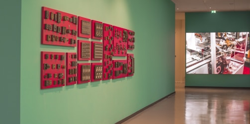 A gallery hallway featuring an art installation on the left with red-framed objects displayed on a teal wall. The installation consists of several red panels with numerous small, vintage-looking items attached, possibly technological or industrial components. On the right, a large digital screen displays an image of machinery with metallic and red elements visible.