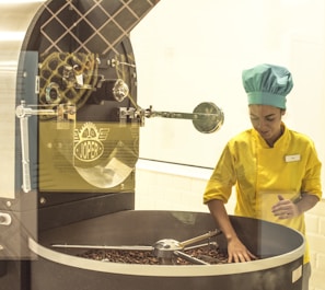A person wearing a yellow uniform and a blue chef's hat is operating a large industrial machine used for roasting coffee beans. The person is inspecting a batch of beans inside a circular roasting drum. The machine is metallic and has the brand 'JOPER' displayed prominently on it.