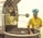 A person wearing a yellow uniform and a blue chef's hat is operating a large industrial machine used for roasting coffee beans. The person is inspecting a batch of beans inside a circular roasting drum. The machine is metallic and has the brand 'JOPER' displayed prominently on it.