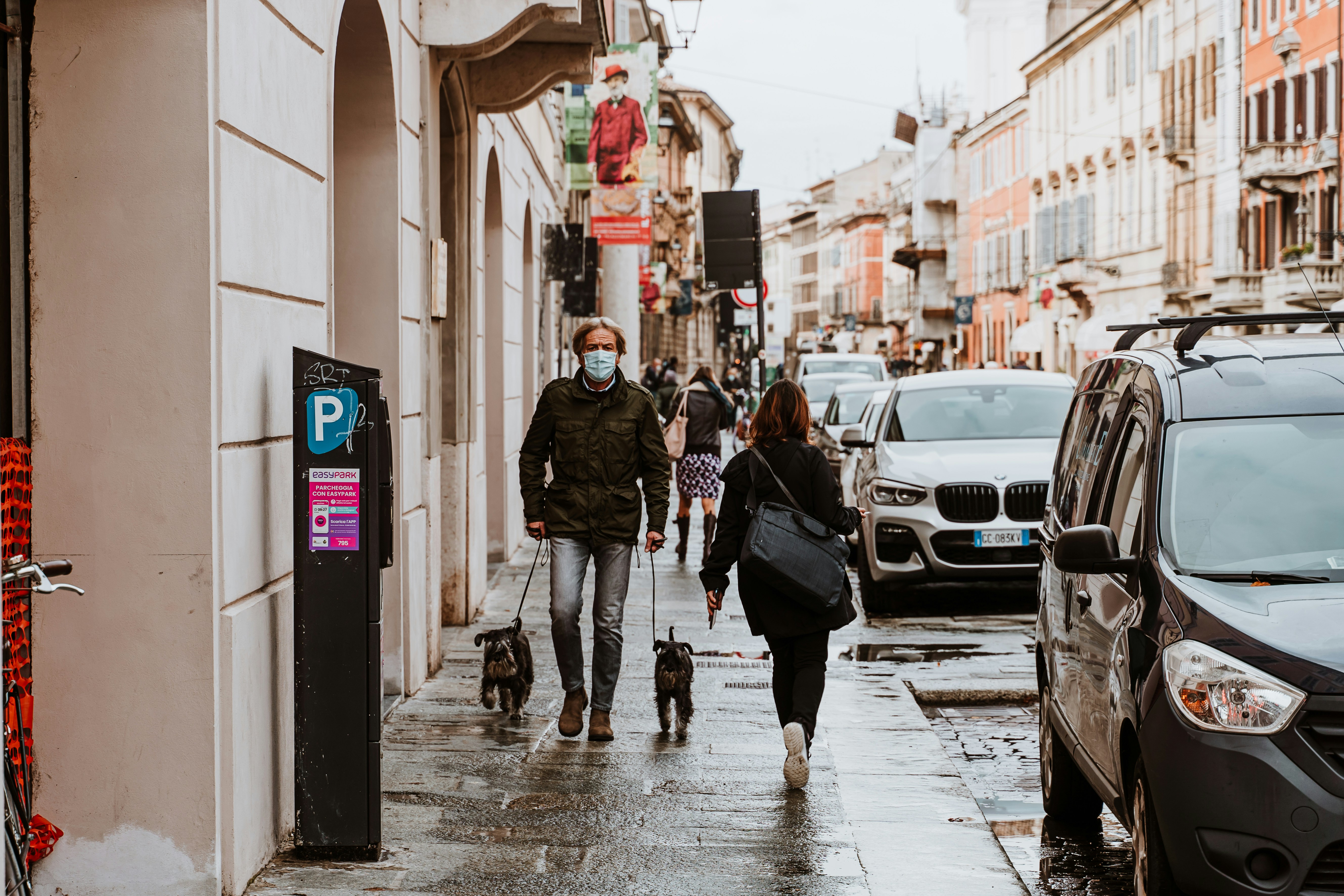 woman in black jacket walking on sidewalk during daytime, A middle aged man wearing a face mask walks his dogs in the city of Parma, Italy, during the coronavirus pandemic
