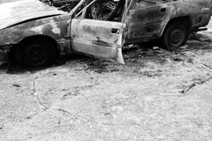 A burned-out car with severe damage sitting on a cracked pavement. The car door is open, showing the interior destruction. The surrounding area includes scattered debris.