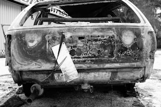 A burnt-out, rusted car with a missing rear window and a detached license plate hanging from a wire. The car's damaged frame displays exposed metal and signs of intense heat.