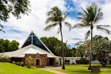 A church with a triangular, modern design roof surrounded by lush greenery and palm trees. The building features a mix of stone and glass elements and is set against a bright, partly cloudy sky.