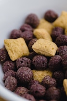 A close-up view of a mix of cereal pieces, including light brown pillow-shaped pieces and dark purple spherical puffs.