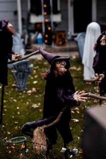 Several Halloween decorations are set up on a lawn, including witch figurines wearing dark robes and pointy hats, holding broomsticks, along with other spooky figures like a ghost figure in the background. The scene is set in front of a house, with other yard decorations visible.
