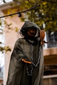 A Halloween decoration features a ghostly figure hanging from a tree branch. The figure wears a dark green, tattered cloak and has a carved pumpkin head with a menacing expression. Chains hang loosely from its body, enhancing the eerie atmosphere. The background shows an out-of-focus building and tree branches with autumn leaves.
