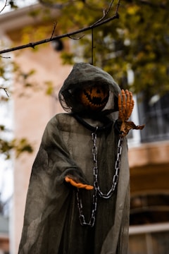 A Halloween decoration features a ghostly figure hanging from a tree branch. The figure wears a dark green, tattered cloak and has a carved pumpkin head with a menacing expression. Chains hang loosely from its body, enhancing the eerie atmosphere. The background shows an out-of-focus building and tree branches with autumn leaves.