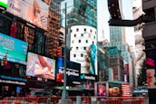 A bustling urban scene features a myriad of colorful advertisements on buildings in a lively city setting. Skyscrapers adorned with bright digital billboards display various advertisements and branding. Traffic signals and vehicles are present in the foreground, with people visible around the area.