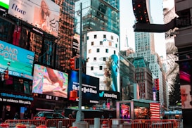 A bustling urban scene features a myriad of colorful advertisements on buildings in a lively city setting. Skyscrapers adorned with bright digital billboards display various advertisements and branding. Traffic signals and vehicles are present in the foreground, with people visible around the area.