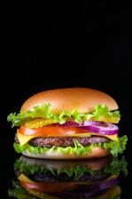 A close-up of a hamburger with a glossy top bun, layers of fresh green lettuce, bright yellow cheese, a juicy beef patty, slices of red tomato, purple onion rings, and sauces, against a black background creating a dramatic contrast.