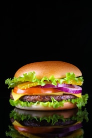 A close-up of a hamburger with a glossy top bun, layers of fresh green lettuce, bright yellow cheese, a juicy beef patty, slices of red tomato, purple onion rings, and sauces, against a black background creating a dramatic contrast.
