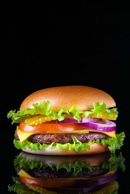 A close-up of a hamburger with a glossy top bun, layers of fresh green lettuce, bright yellow cheese, a juicy beef patty, slices of red tomato, purple onion rings, and sauces, against a black background creating a dramatic contrast.