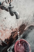 A metal water tap is visibly aged and slightly rusted, with water flowing steadily from its spout into a red bucket below. The background features a textured, weathered wall with patches of discoloration.