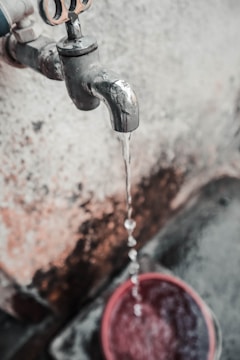 A metal water tap is visibly aged and slightly rusted, with water flowing steadily from its spout into a red bucket below. The background features a textured, weathered wall with patches of discoloration.