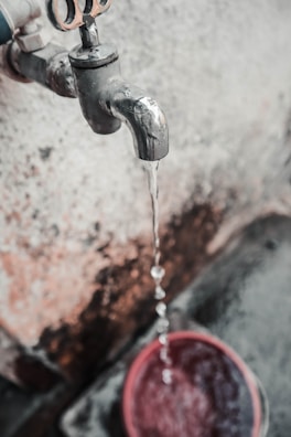 A metal water tap is visibly aged and slightly rusted, with water flowing steadily from its spout into a red bucket below. The background features a textured, weathered wall with patches of discoloration.