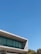 A modernist building with large glass windows and concrete structure under a clear blue sky. The building features clean lines and a minimalistic design. Trees with sparse branches appear at the bottom of the image, alongside greenery in the background.