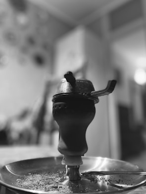 A metal hookah or shisha pipe is situated against a blurred indoor background. The focus is on the top part of the hookah, with intricate details of the coal tray and bowl visible. The environment appears domestic with subtle shadows and light creating a calm atmosphere.
