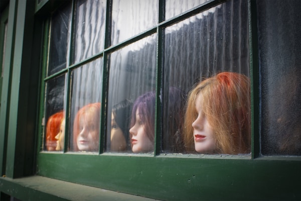 Several mannequin heads with colorful wigs are displayed in the window of a green-framed storefront. The wigs vary in vibrant colors, including orange, red, and purple, and the mannequins are positioned in a row, each facing a different direction.