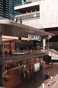A modern, multi-story building with large glass windows displaying the Huawei logo. People are walking on the upper and lower levels, some inside the store, and others outside on the street. Reflections and various electronic products are visible through the large windows, suggesting a retail environment. The surrounding architecture is sleek and contemporary.