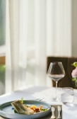 A neatly set dining table with a plate of gourmet food, focusing on a beautifully plated dish that includes a piece of fish, vegetables, and garnishes. The table also holds a wine glass, a water glass, and cutlery, all arranged on a placemat. Soft, natural light filters in through sheer curtains in the background, creating a serene and elegant ambiance.