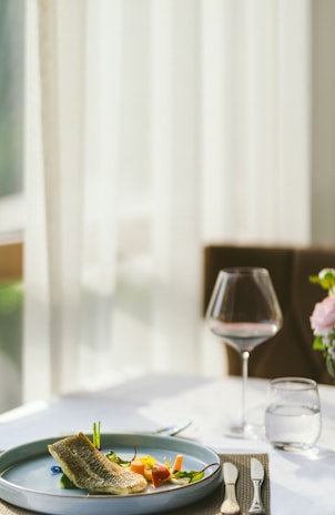 A neatly set dining table with a plate of gourmet food, focusing on a beautifully plated dish that includes a piece of fish, vegetables, and garnishes. The table also holds a wine glass, a water glass, and cutlery, all arranged on a placemat. Soft, natural light filters in through sheer curtains in the background, creating a serene and elegant ambiance.