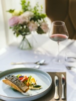 A sophisticated dining setting features a carefully plated dish with seared fish and colorful vegetables on a white plate. A glass of red wine is nearby, set on a white tablecloth with a brown woven placemat. In the background, there's a soft focus arrangement of pastel flowers.