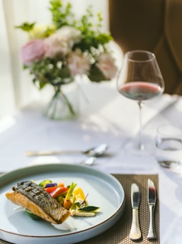 A sophisticated dining setting features a carefully plated dish with seared fish and colorful vegetables on a white plate. A glass of red wine is nearby, set on a white tablecloth with a brown woven placemat. In the background, there's a soft focus arrangement of pastel flowers.