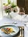 A sophisticated dining setting features a carefully plated dish with seared fish and colorful vegetables on a white plate. A glass of red wine is nearby, set on a white tablecloth with a brown woven placemat. In the background, there's a soft focus arrangement of pastel flowers.