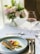 A sophisticated dining setting features a carefully plated dish with seared fish and colorful vegetables on a white plate. A glass of red wine is nearby, set on a white tablecloth with a brown woven placemat. In the background, there's a soft focus arrangement of pastel flowers.