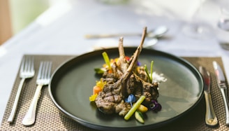 Three lamb chops are elegantly arranged on a black plate, garnished with a variety of vibrant vegetables and edible flowers. The setting is a refined table with a woven placemat, accompanied by sophisticated cutlery, suggesting a fine dining experience.