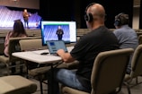 Three people are seated at a table in an auditorium, wearing headphones and using a computer setup with dual monitors to livestream or manage a recording. One person is using a laptop, and the others are monitoring the screen. In the background, a speaker stands on the stage, addressing an audience with a guitar visible next to them. The auditorium is largely empty with rows of beige seats.