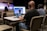 Three people are seated at a table in an auditorium, wearing headphones and using a computer setup with dual monitors to livestream or manage a recording. One person is using a laptop, and the others are monitoring the screen. In the background, a speaker stands on the stage, addressing an audience with a guitar visible next to them. The auditorium is largely empty with rows of beige seats.
