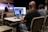 Three people are seated at a table in an auditorium, wearing headphones and using a computer setup with dual monitors to livestream or manage a recording. One person is using a laptop, and the others are monitoring the screen. In the background, a speaker stands on the stage, addressing an audience with a guitar visible next to them. The auditorium is largely empty with rows of beige seats.