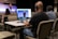 Three people are seated at a table in an auditorium, wearing headphones and using a computer setup with dual monitors to livestream or manage a recording. One person is using a laptop, and the others are monitoring the screen. In the background, a speaker stands on the stage, addressing an audience with a guitar visible next to them. The auditorium is largely empty with rows of beige seats.