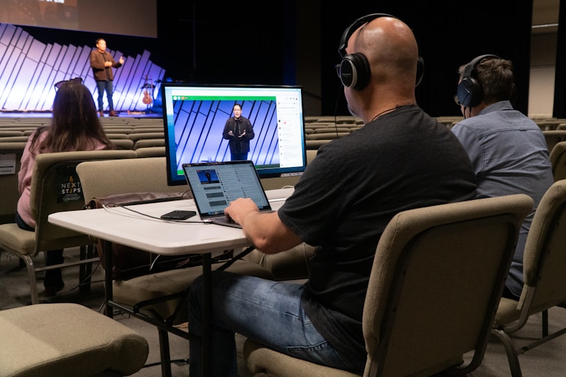 Three people are seated at a table in an auditorium, wearing headphones and using a computer setup with dual monitors to livestream or manage a recording. One person is using a laptop, and the others are monitoring the screen. In the background, a speaker stands on the stage, addressing an audience with a guitar visible next to them. The auditorium is largely empty with rows of beige seats.