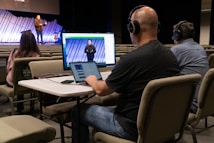 Three people are seated at a table in an auditorium, wearing headphones and using a computer setup with dual monitors to livestream or manage a recording. One person is using a laptop, and the others are monitoring the screen. In the background, a speaker stands on the stage, addressing an audience with a guitar visible next to them. The auditorium is largely empty with rows of beige seats.