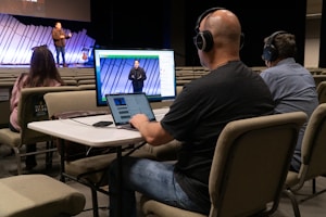 Three people are seated at a table in an auditorium, wearing headphones and using a computer setup with dual monitors to livestream or manage a recording. One person is using a laptop, and the others are monitoring the screen. In the background, a speaker stands on the stage, addressing an audience with a guitar visible next to them. The auditorium is largely empty with rows of beige seats.