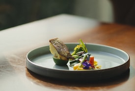 A beautifully arranged dish on a sleek, modern plate features a piece of seared fish accompanied by an assortment of colorful vegetables and edible flowers. The presentation is minimalist, with an emphasis on balance and natural beauty.