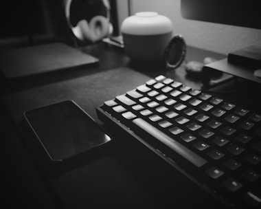 A black and white photo of a minimalist desk setup featuring a mechanical keyboard and a smartphone. The desk is organized with a sleek lamp and a pair of headphones in the background. The lighting creates a soft, moody atmosphere.