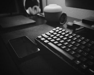 A black and white photo of a minimalist desk setup featuring a mechanical keyboard and a smartphone. The desk is organized with a sleek lamp and a pair of headphones in the background. The lighting creates a soft, moody atmosphere.