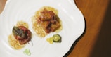 A plate containing a gourmet dish, featuring cubes of glazed meat or tofu in a sauce, garnished with a fresh green leaf. A small slice of citrus fruit and colorful gel-like spheres are also present on the plate, all arranged artistically on a white, wavy-edged dish.