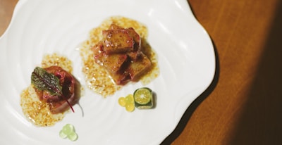 A plate containing a gourmet dish, featuring cubes of glazed meat or tofu in a sauce, garnished with a fresh green leaf. A small slice of citrus fruit and colorful gel-like spheres are also present on the plate, all arranged artistically on a white, wavy-edged dish.