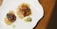 A plate containing a gourmet dish, featuring cubes of glazed meat or tofu in a sauce, garnished with a fresh green leaf. A small slice of citrus fruit and colorful gel-like spheres are also present on the plate, all arranged artistically on a white, wavy-edged dish.