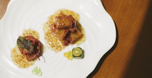 A plate containing a gourmet dish, featuring cubes of glazed meat or tofu in a sauce, garnished with a fresh green leaf. A small slice of citrus fruit and colorful gel-like spheres are also present on the plate, all arranged artistically on a white, wavy-edged dish.