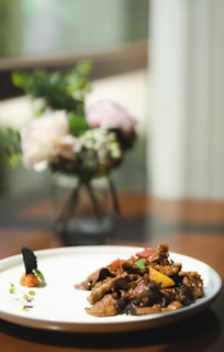 A white plate with an artful arrangement of finely cooked meat pieces and colorful vegetables, placed on a polished wooden table. The background features a soft focus on a glass vase holding a bouquet of delicate flowers in pastel shades.