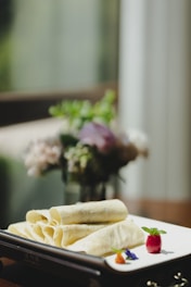 Delicate crepes are neatly arranged on a white plate, garnished with a small, vibrant parfait and colorful flowers. A blurred background shows soft light filtering through a window, with subtle hints of green and purple from out-of-focus plants or flowers.