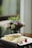 Delicate crepes are neatly arranged on a white plate, garnished with a small, vibrant parfait and colorful flowers. A blurred background shows soft light filtering through a window, with subtle hints of green and purple from out-of-focus plants or flowers.